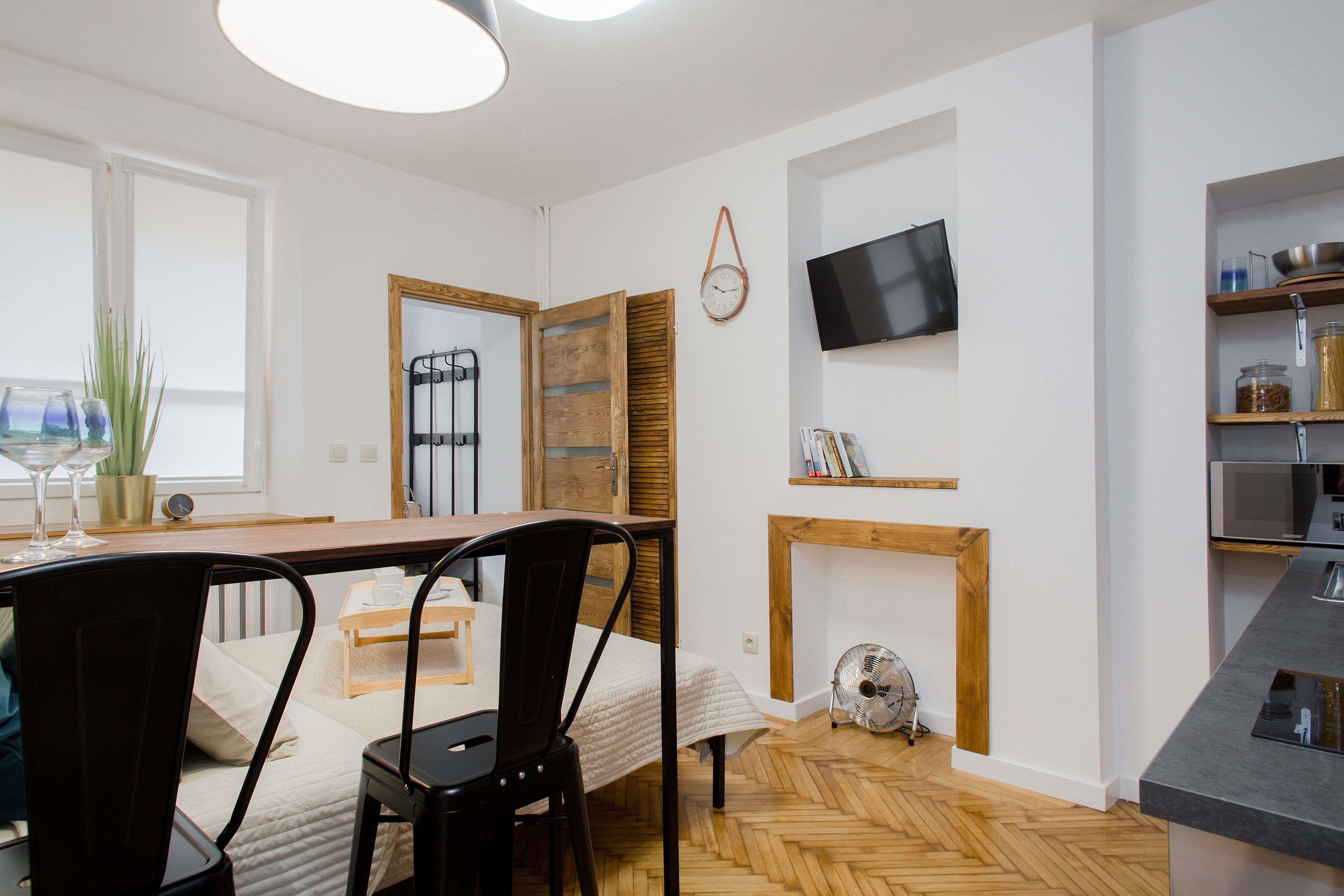 A modern kitchen with a wooden dining table, chairs, a TV mounted on the wall, and a built-in oven.