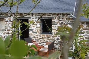 A stone building with a sloped roof, a patio area with wooden furniture, and a view of the surrounding greenery. - La Vieille Ferme (Landevennec)