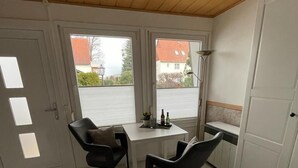 A compact room with a wooden ceiling, a small table with two chairs, a window with a view of buildings, and a door. - Holiday house in a quiet location and near the forest (Wernigerode)