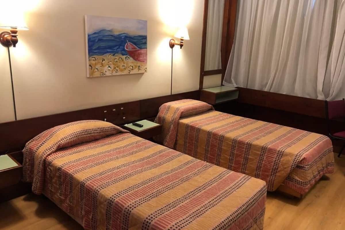 A hotel room with two single beds, a nightstand, a lamp, a painting on the wall, and a window with curtains.