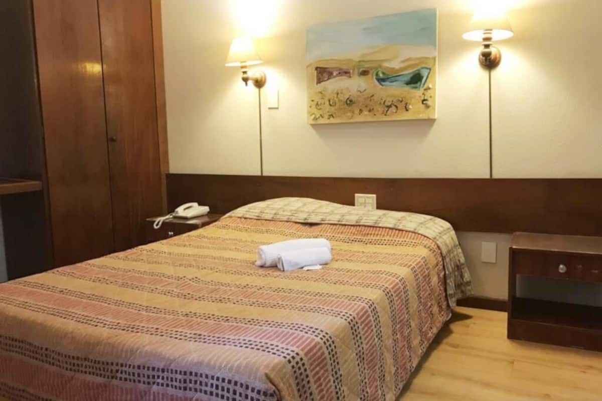 A hotel room with a bed, a bedside telephone, a painting on the wall, and two wall-mounted lamps.