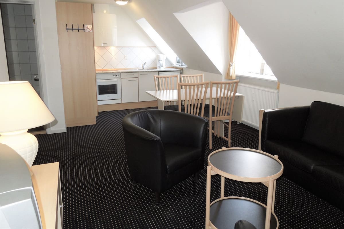Een compacte hotelkamer met een kitchenette, een eetgedeelte en een zithoek met een zwarte leren fauteuil en een klein rond tafeltje.