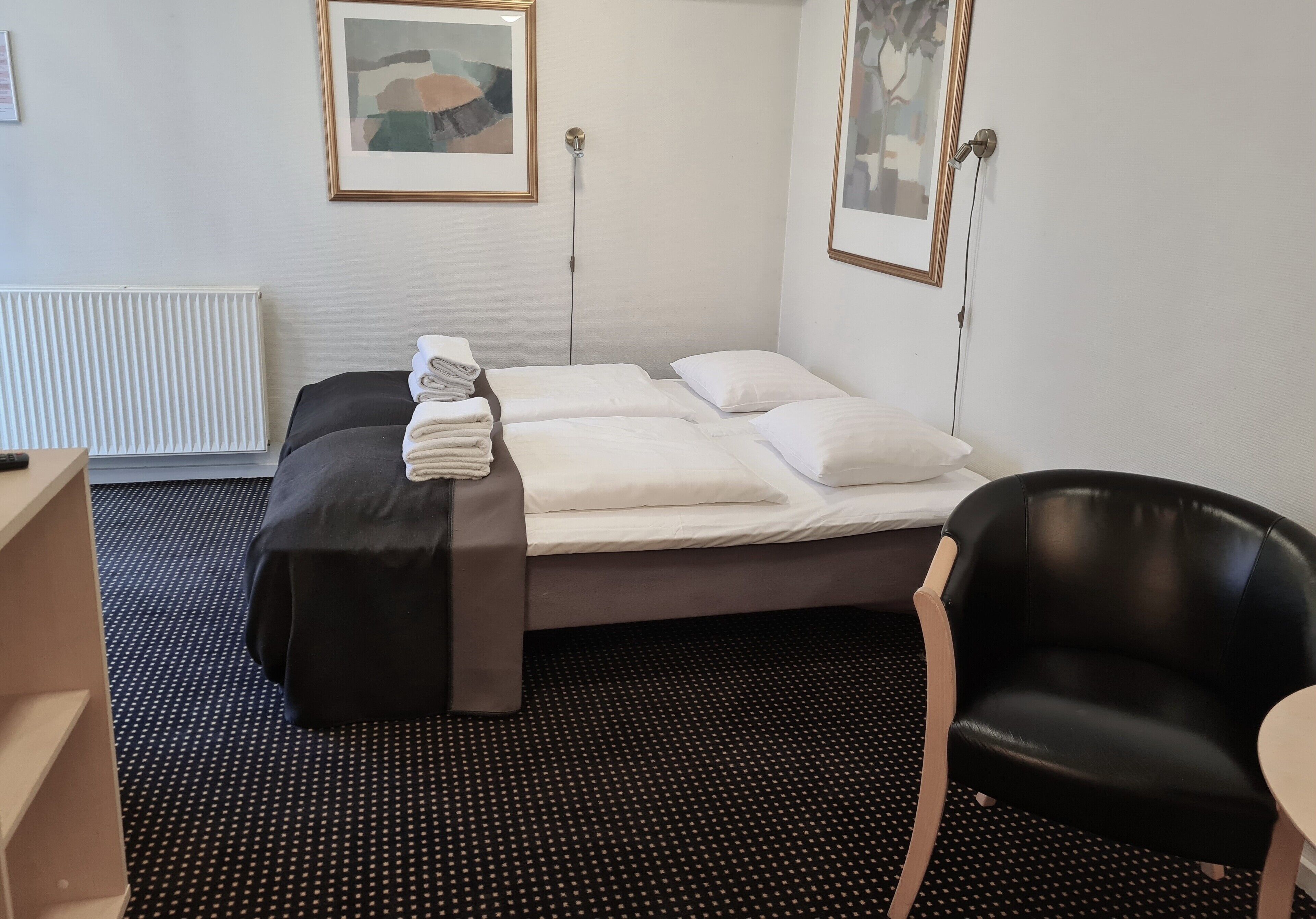 A hotel room with a bed, a black chair, a small table, and artwork on the walls.