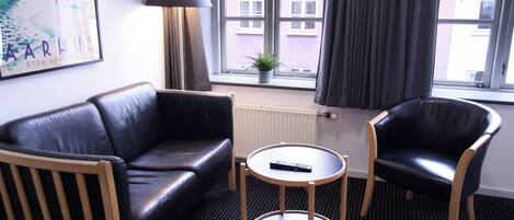 Ein Hotelzimmer mit einer schwarzen Ledersofa, einem Holzstuhl, einem kleinen Rundtisch und einem Fenster mit Vorhängen.