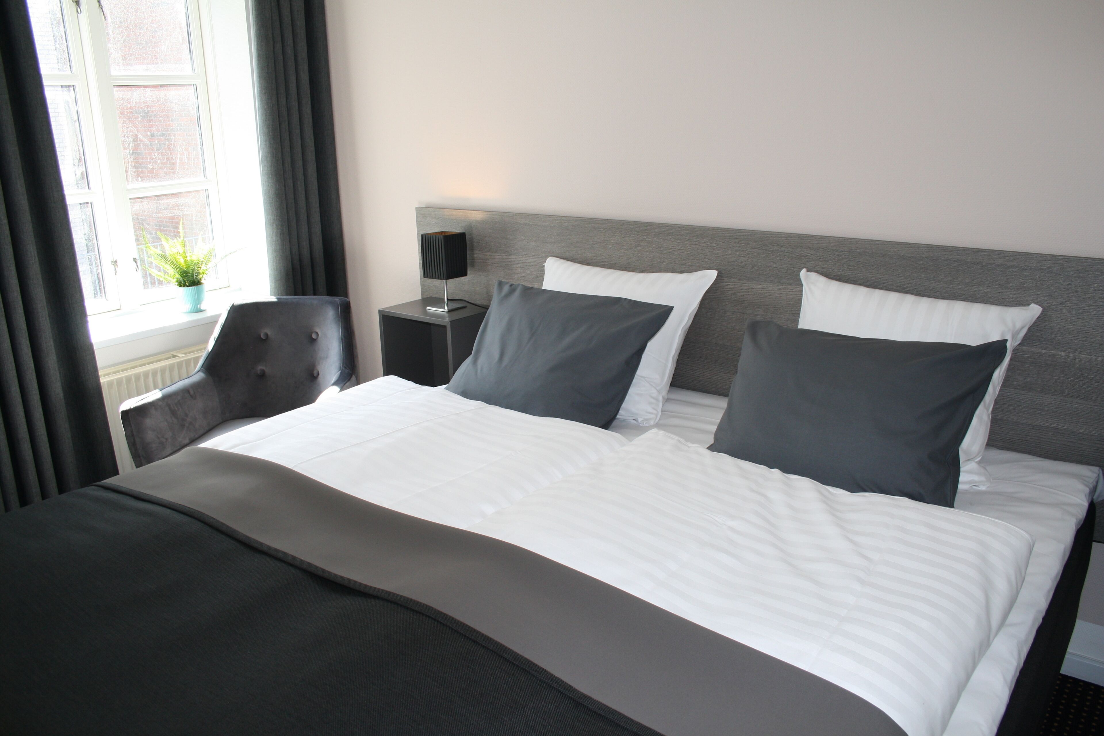 Una cama bien hecha con ropa de cama blanca y gris, un cabecero gris y una mesita de noche con una lámpara.