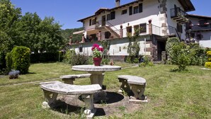 Une maison à deux étages avec un jardin agrémenté d’une table et de bancs en pierre, le tout entouré de verdure et de fleurs.
