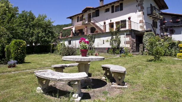 Una casa de dos pisos con jardín, que incluye una mesa de piedra y bancos, rodeada de follaje y flores.