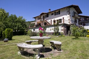 Una casa de dos pisos con jardín, que incluye una mesa de piedra y bancos, rodeada de follaje y flores.