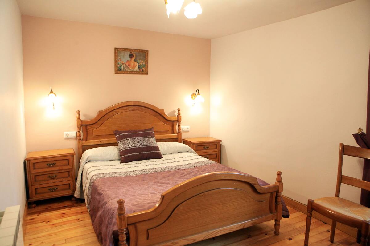 Quarto com cama de madeira, duas mesas de cabeceira, um criado-mudo, uma cadeira e um quadro na parede.