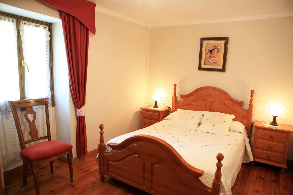 Quarto com cama de madeira, duas mesas de cabeceira com luminárias, uma cadeira e janela com cortinas.