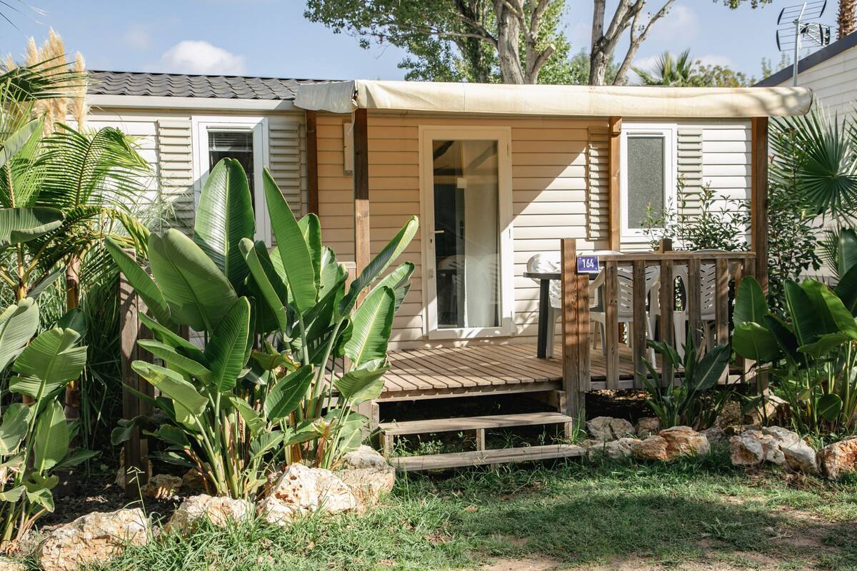 Eenlaags huis met een houten terras, omgeven door tropische planten en een pad van stenen.