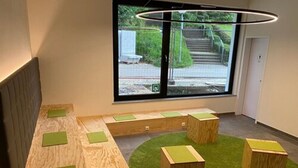 A modern room with a long wooden bench, a small green carpet, and a window with a view of a staircase and greenery outside. - a2 Hotels Wernau (Wernau)