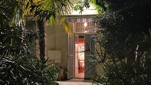 Ein Weg führt zu einem Gebäude mit Balkon, umgeben von dichtem Grün und einem gut beleuchteten Eingang.