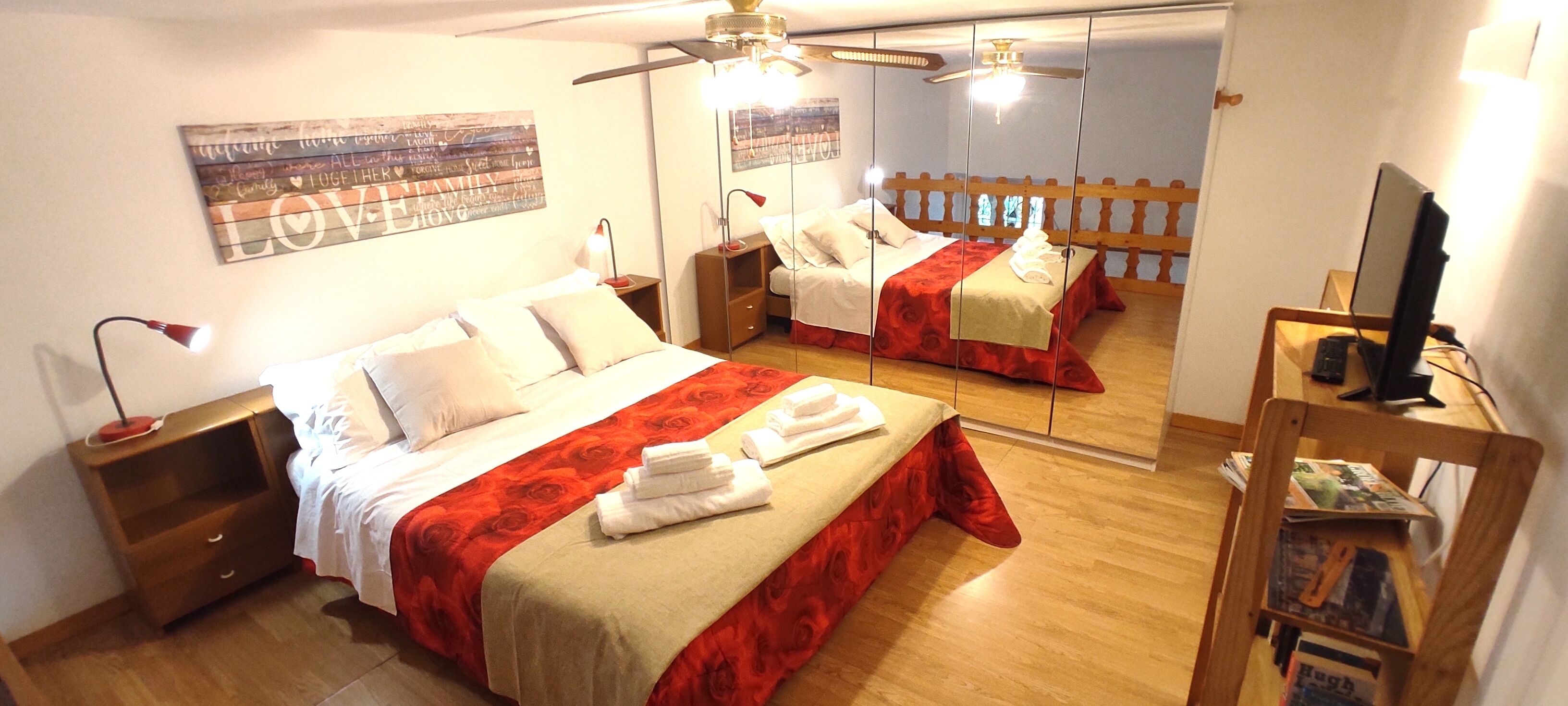 A well-lit bedroom with a large bed, a bedside table, a mirror, and a television on a wooden stand.