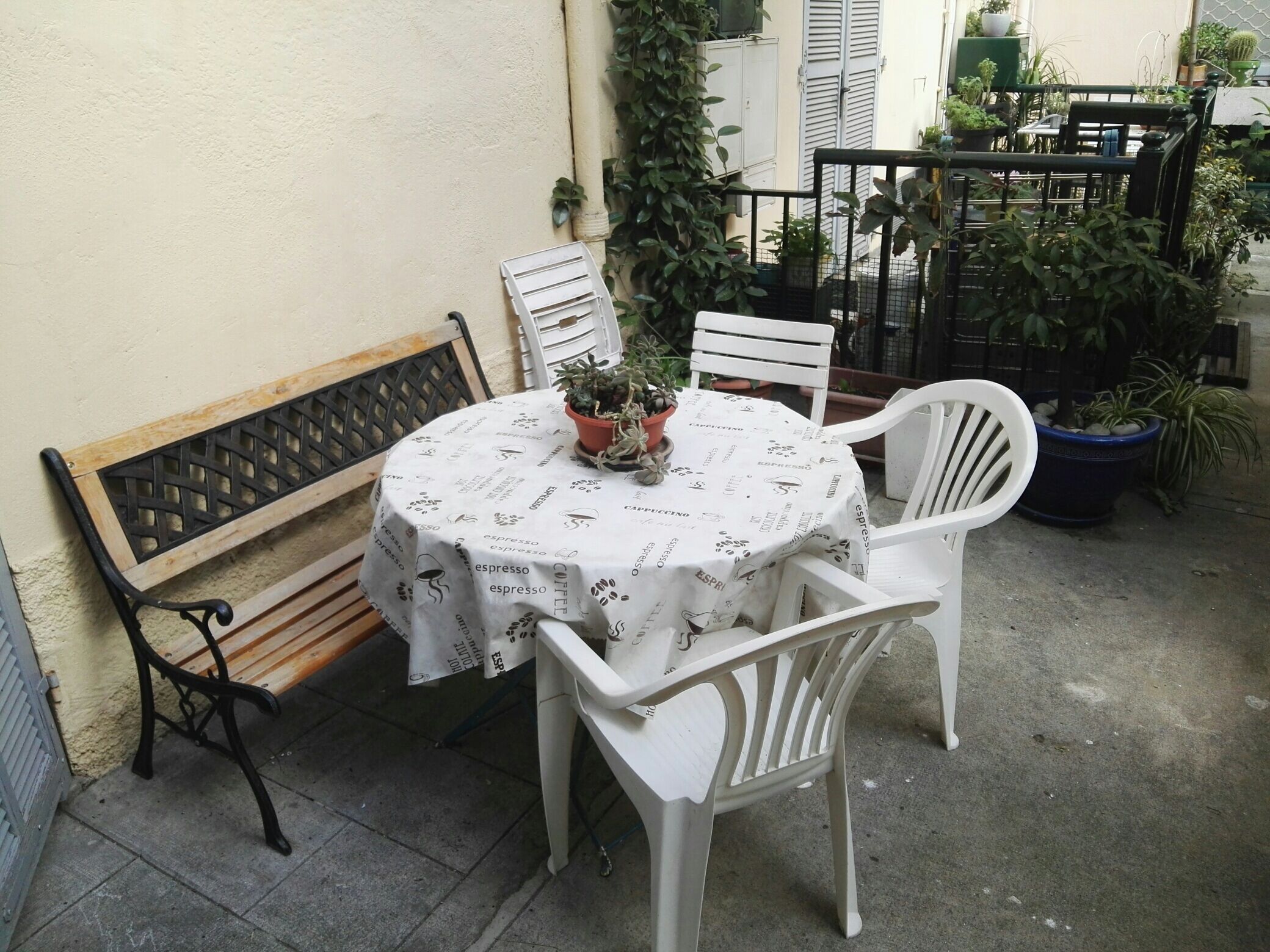A small outdoor seating area with a round table covered by a white tablecloth with espresso-related text, surrounded by white plastic chairs.