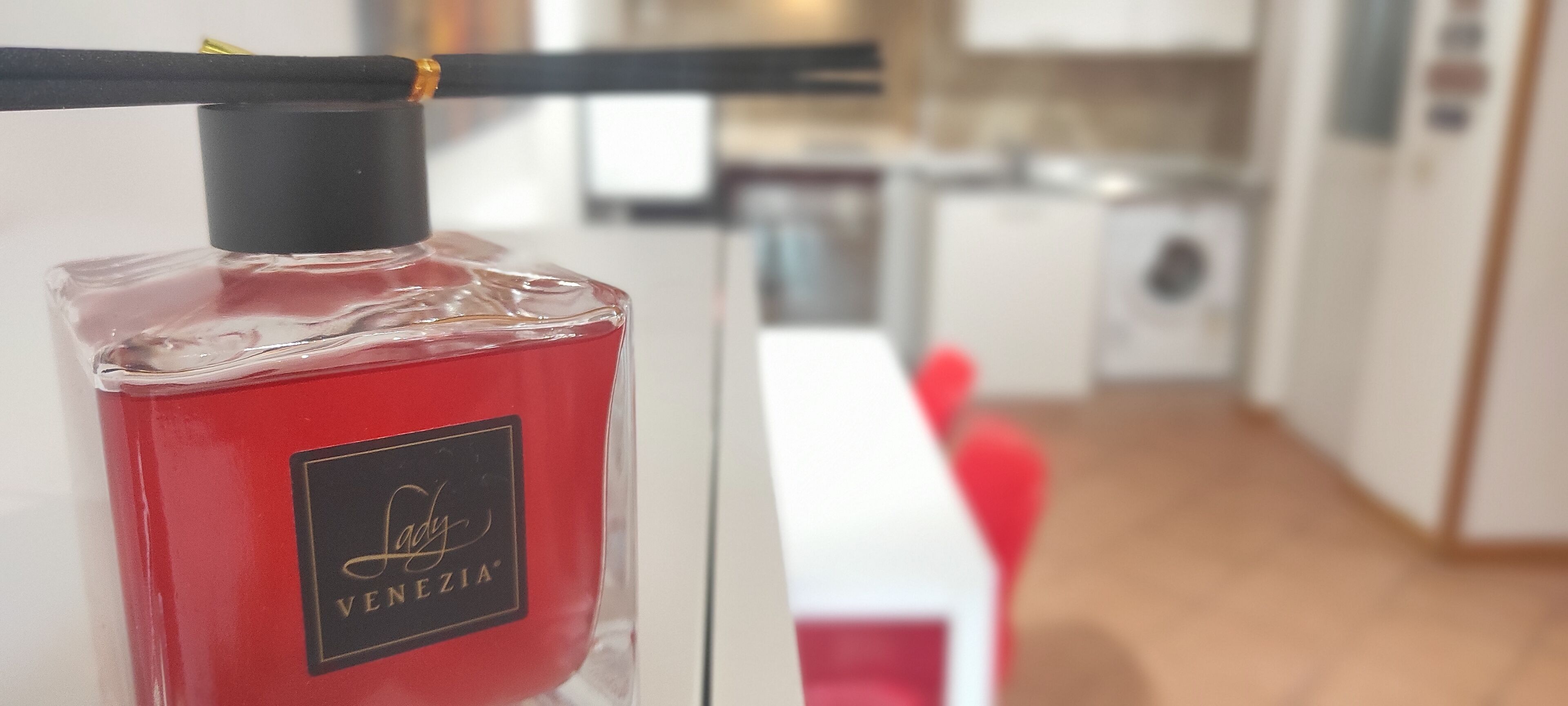 A bottle of Lady Venezia perfume with a black cap and a gold label, placed on a white surface in a modern kitchen with white cabinets and a wooden floor.