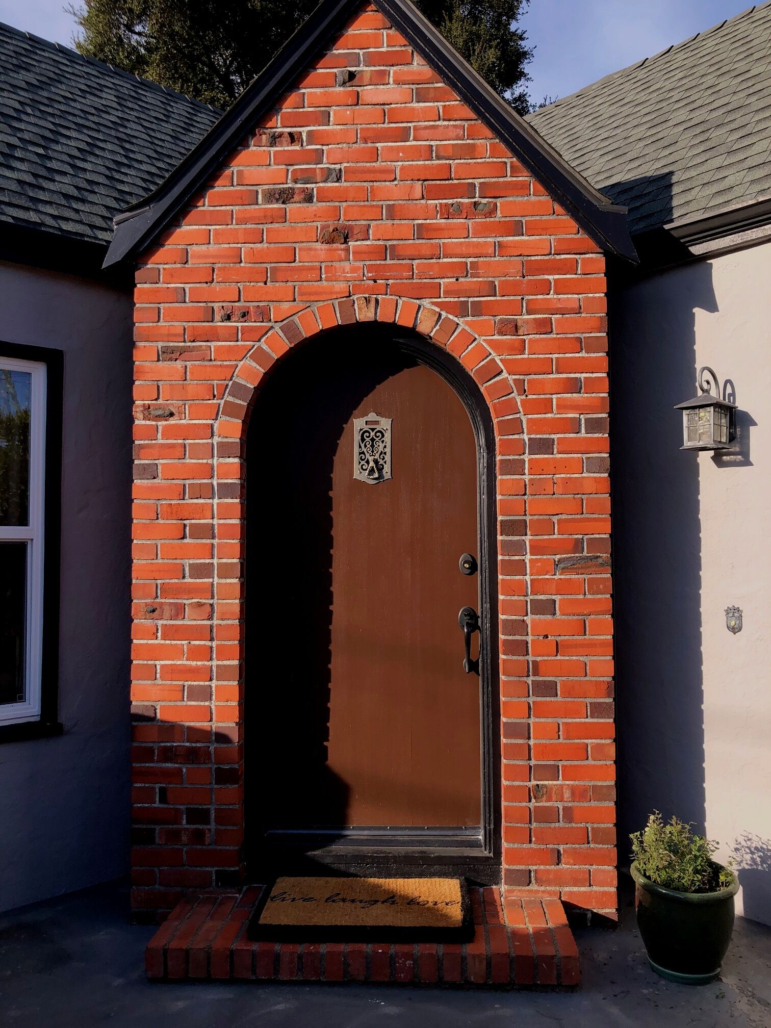 A brick entrance with a wooden door, a doormat, and a potted plant.