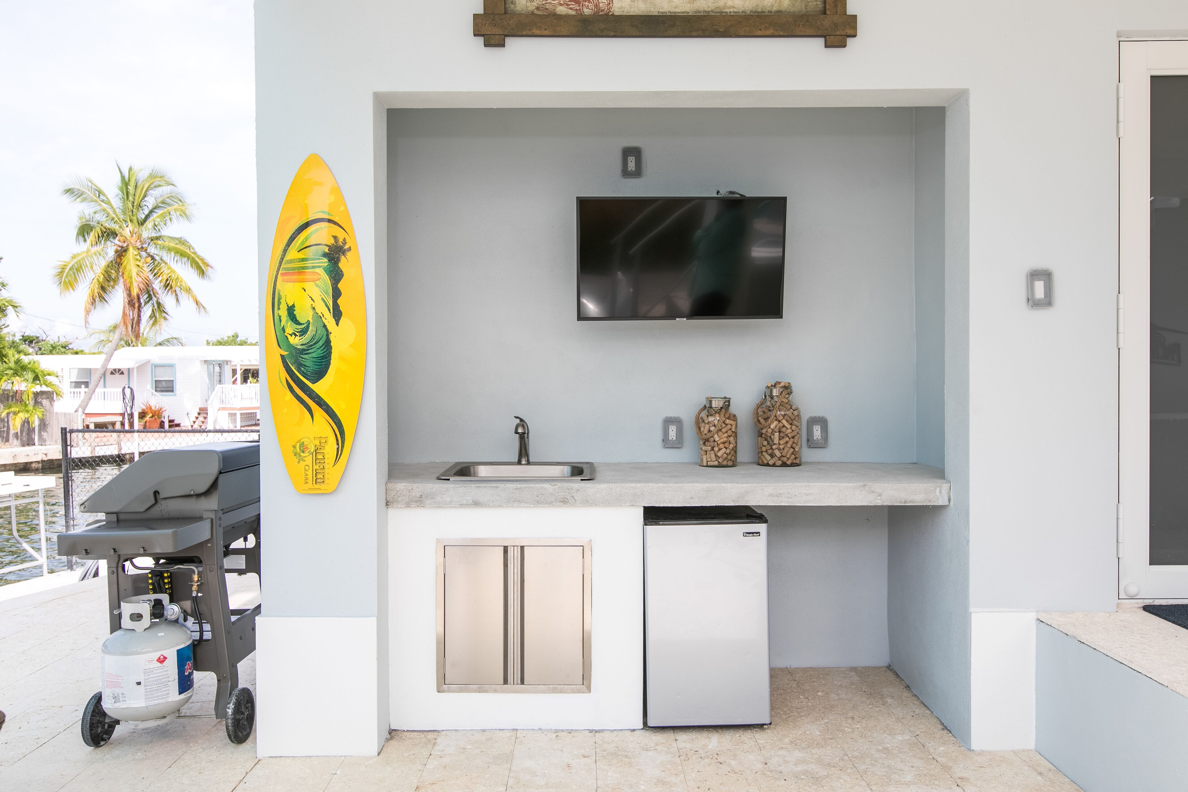 Une cuisine compacte équipée d’un lave-vaisselle intégré, d’un téléviseur à écran plat fixé au-dessus de l’évier et d’une décoration murale représentant une planche de surf.