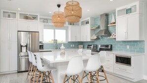 Una cocina moderna con armarios blancos, un refrigerador de acero inoxidable, una isla grande con sillas blancas y un salpicadero de azulejos azules.