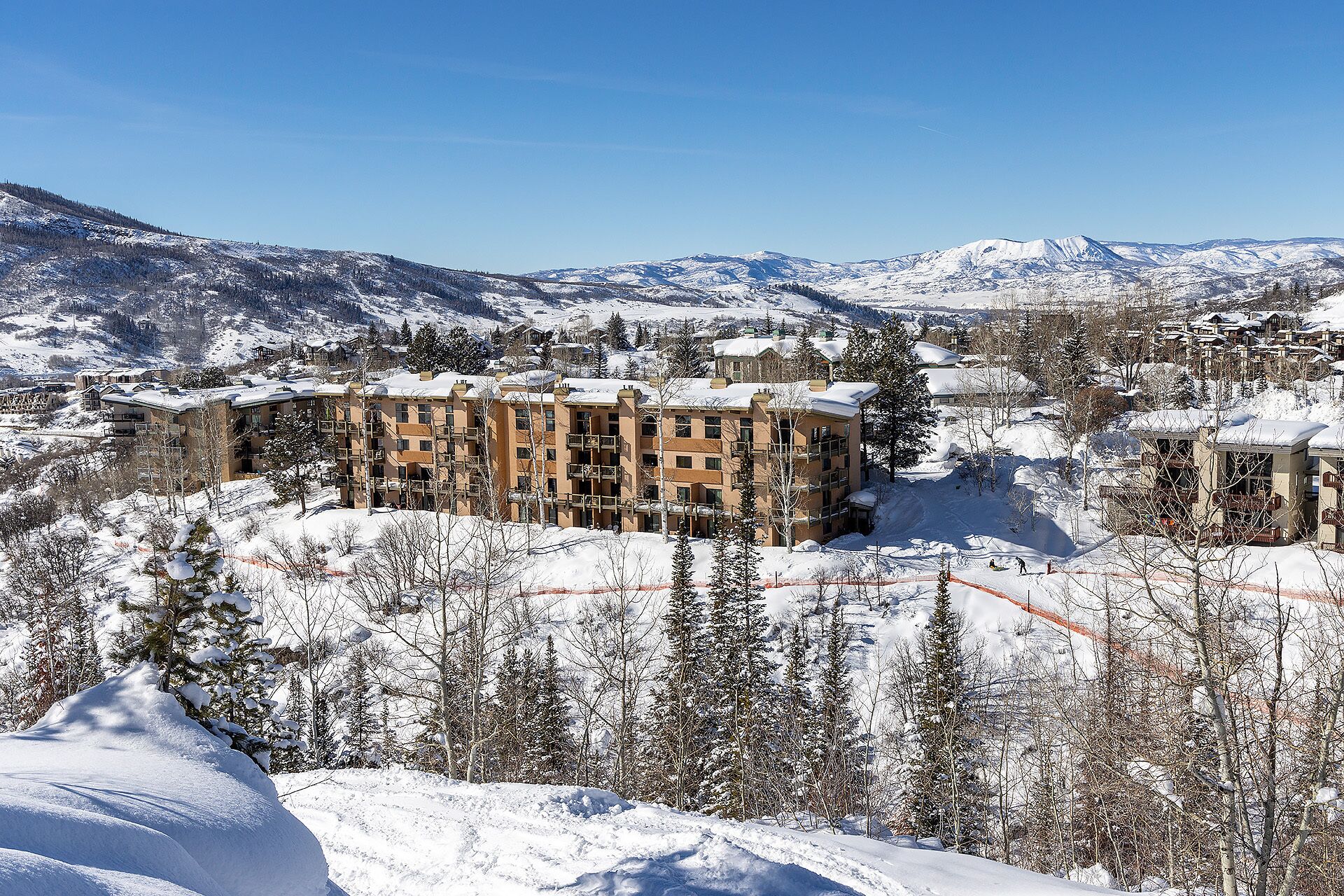 A snow-covered mountain resort with multi-story buildings, ski slopes, and a clear blue sky.