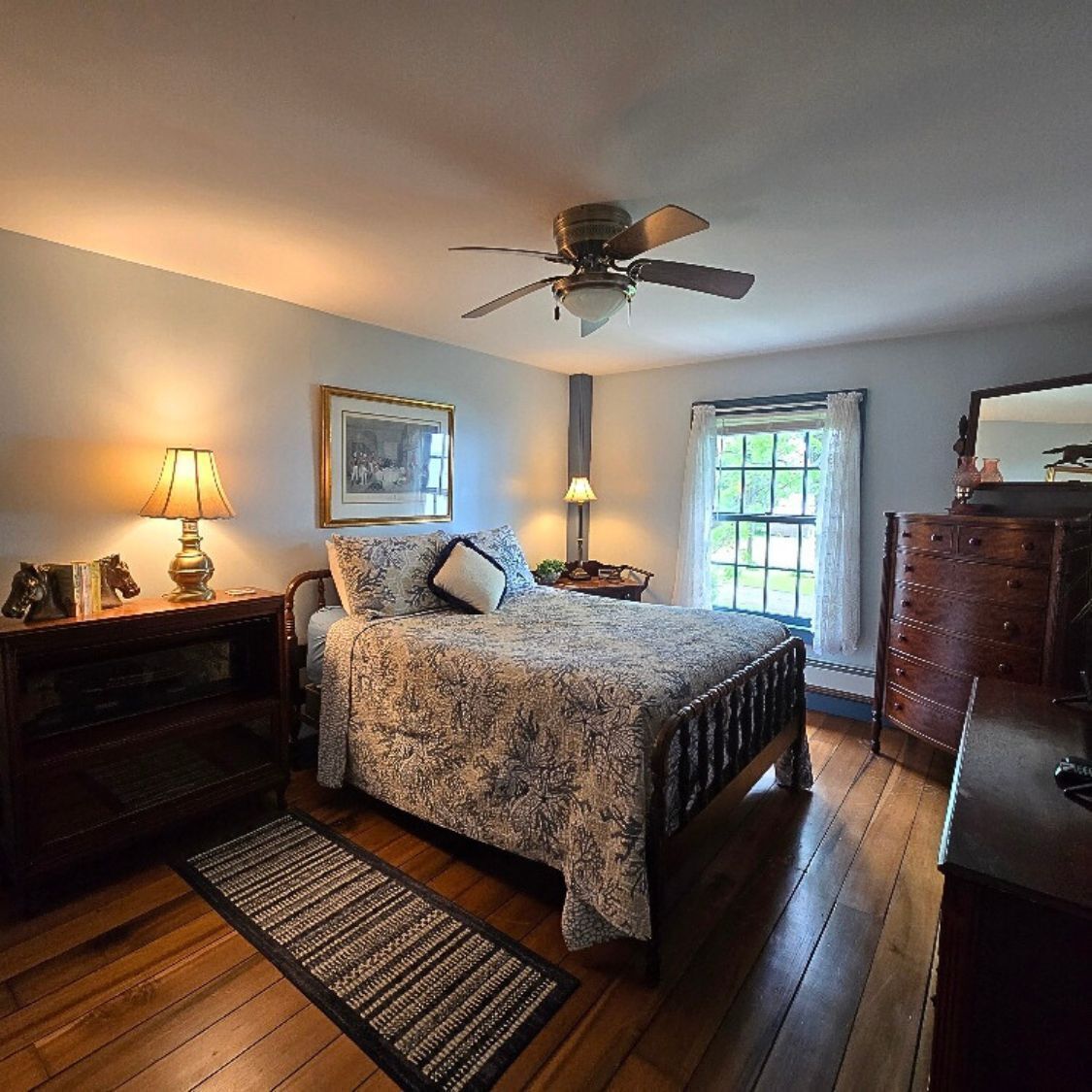 Une chambre à coucher comprenant un lit, un ventilateur de plafond, une commode, une table de chevet, une lampe, un tableau encadré et une fenêtre avec des rideaux.