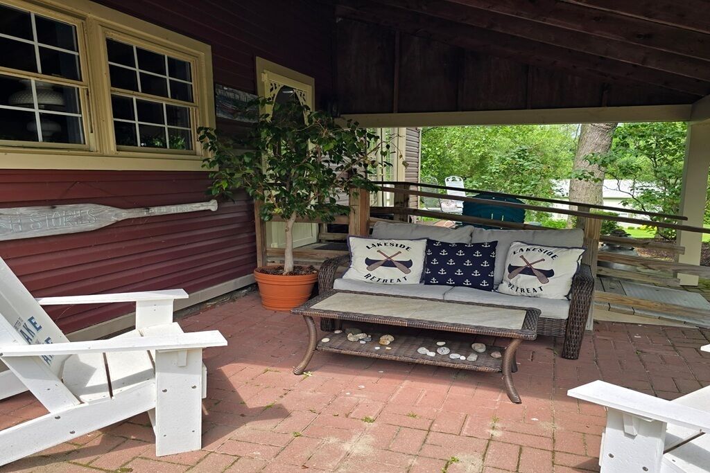 Une terrasse couverte avec un banc en osier, une plante en pot et une chaise Adirondack blanche.