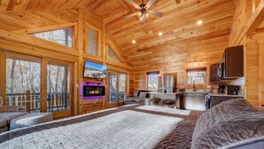 A log cabin interior with a large bed, a television, a kitchen area, and a balcony with a view of trees. - Cozy Rose` cabin for up to 6 guests - Hocking Hills - Hot Tub-Fire Pit-Wifi (Logan)