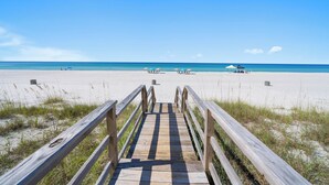A wooden boardwalk leading to a beach with clear blue water and a few beach umbrellas.