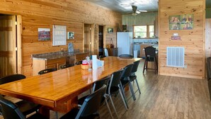 A wooden interior with a long dining table, chairs, a kitchen area, and a ceiling fan.
