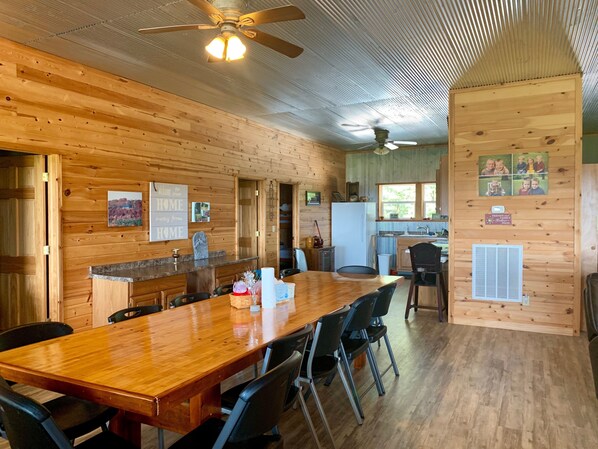 A wooden interior with a long dining table, chairs, a kitchen area, and a ceiling fan.