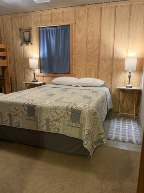 A bedroom with a large bed, two bedside tables with lamps, a wooden headboard, a window with a blue curtain, and a small wooden chair.