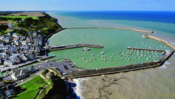 An aerial view of a coastal town with a marina, a lighthouse, and a historic stone building. - Large bright house in the shape of a boat (Port-en-Bessin-Huppain)