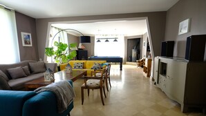 A living room with a sofa, a dining table, chairs, a coffee table, a plant, and a kitchen area with cabinets and a stove. - Large bright house in the shape of a boat (Port-en-Bessin-Huppain)