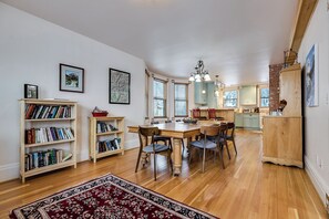 A spacious living area with a wooden dining table surrounded by chairs, a bookshelf, and a large rug.