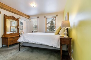 Een slaapkamer met een bed, een dressoir, een spiegel, een lamp en een raam met uitzicht op een besneeerd landschap.