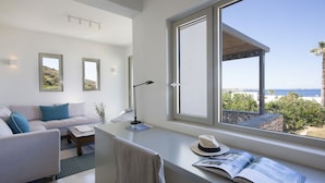 A modern living room with a large window, a sofa, a coffee table, and a desk with a hat and book. - Villa Rinia - Seaview villa (Ampelas)
