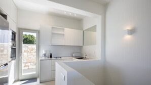 A modern kitchen with white cabinetry, a built-in oven, and a stone wall visible through the door. - Villa Rinia - Seaview villa (Ampelas)