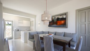 A modern kitchen with white cabinetry, a dining area with a grey sofa and chairs, and a painting above the sofa. - Villa Rinia - Seaview villa (Ampelas)