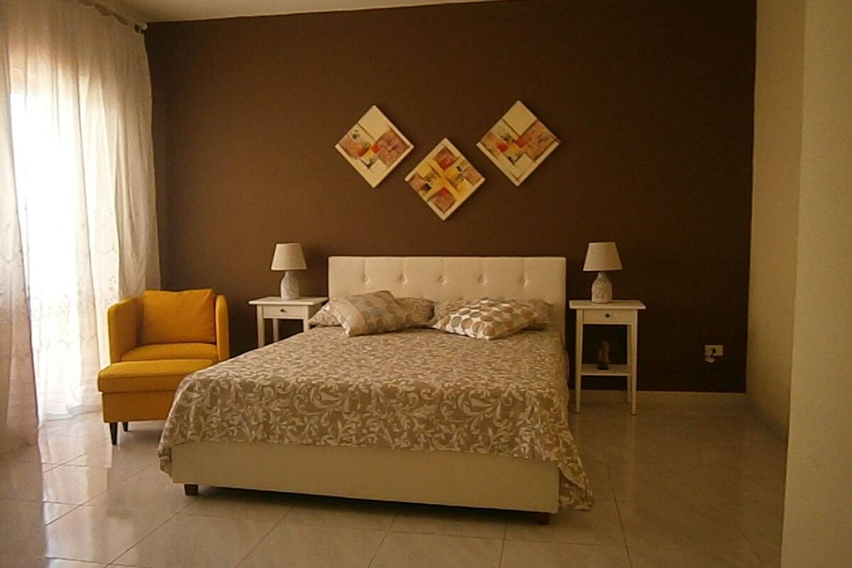 Un dormitorio con una cama, dos mesitas de noche con lámparas, un sillón amarillo y dos obras de arte colgadas en la pared.