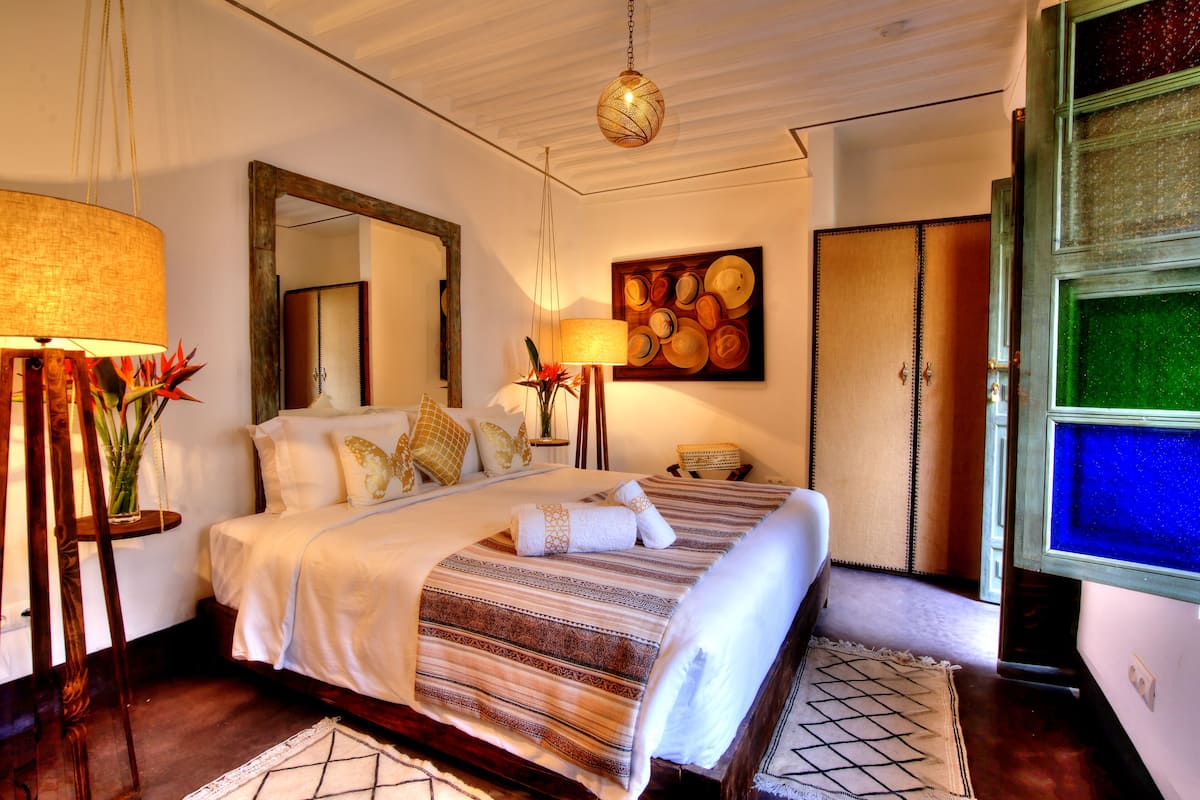 A well-lit bedroom with a large bed, a mirror, a wall art piece, and a green-tinted window.