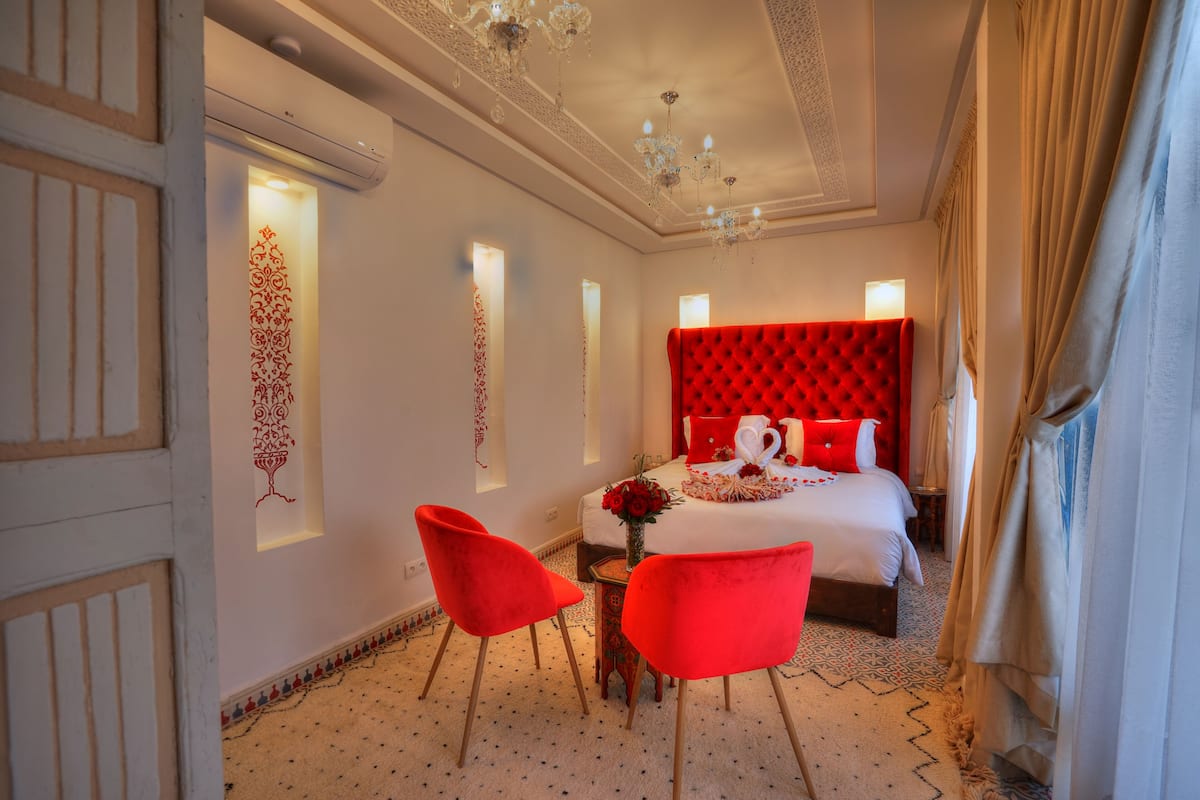 A hotel room with a red upholstered headboard, a large bed with white linens, two red chairs, and a small table with a vase of flowers.