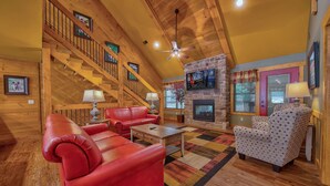 A spacious living room with a fireplace, red leather sofas, a patterned armchair, wooden flooring, a staircase, and a ceiling fan.