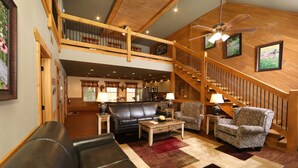 A spacious living area with a black leather sofa, patterned armchairs, and a wooden coffee table. A staircase with a wooden railing leads to an upper level. - Head Over Hills (Gatlinburg)
