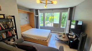 A modern hotel room with a bed, a sofa, a TV, a bookshelf, and large windows with a view of greenery. - Modern, clean & private 1BR suite. Up to 4 guests. (Surrey)