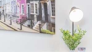 Une pièce comprenant un banc recouvert d’un tissu gris, une lampe fixée au mur, une plante en pot et un tableau représentant une scène de rue.