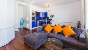 Un salon moderne avec un canapé et un pouf gris, une table basse en verre, et une cuisine en arrière-plan avec des armoires bleues et des plans de travail blancs.