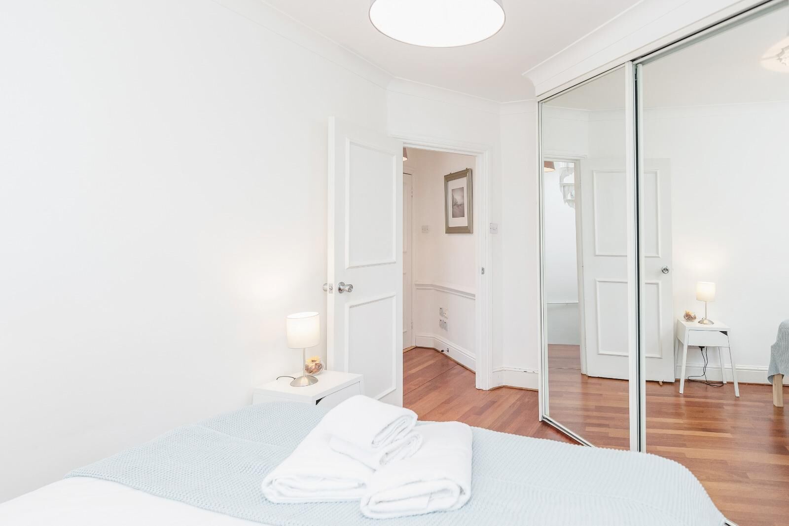 A minimalist bedroom with a large mirror, a neatly made bed, and a small bedside table with a lamp.