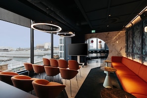 Ein stilvoller Lounge-Bereich mit Blick auf die Stadt, mit orangefarbenen Sitzgelegenheiten, einem kleinen Tisch und einem großen Flachbildfernseher.