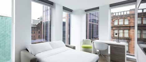 A modern bedroom with a bed, a desk, and a chair, featuring large windows and a view of the city.