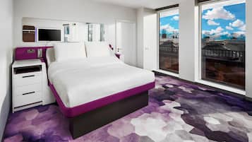 Una habitación de hotel moderna con una cama grande, una mesita de noche, un televisor y una ventana con vistas a la ciudad.